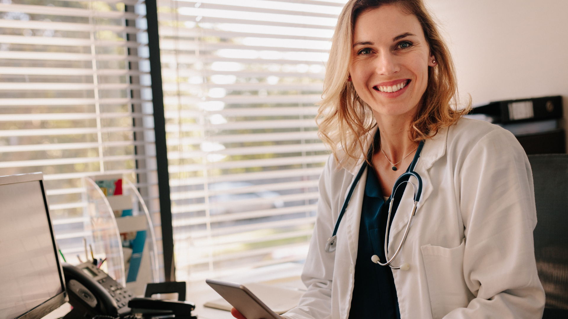woman physician involved in education sat at a desk holding her phone whilst smiling at the camera