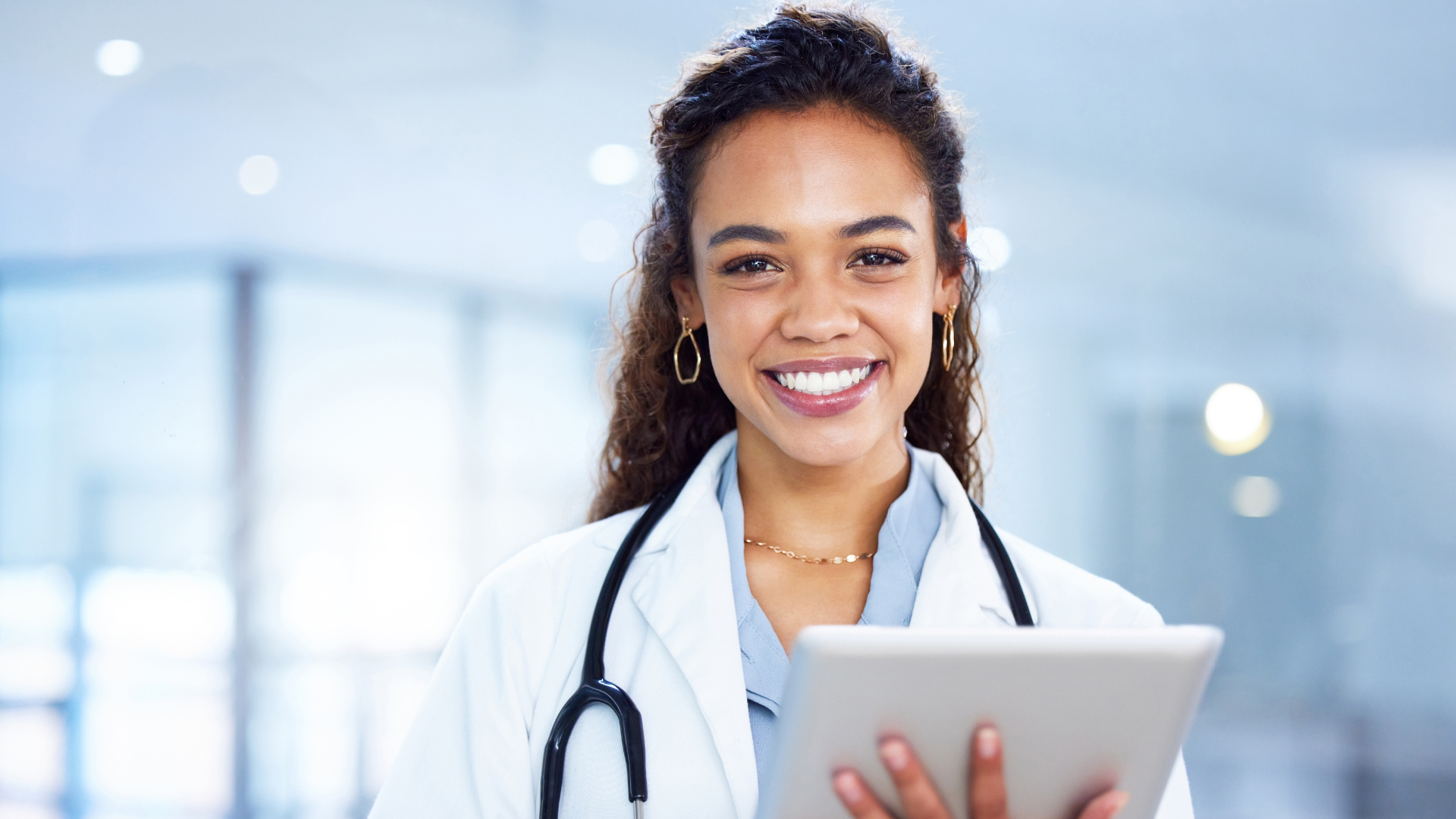 Young woman physician stood in a clinical environment holding an ipad, smiling at the camera, representing women physicians