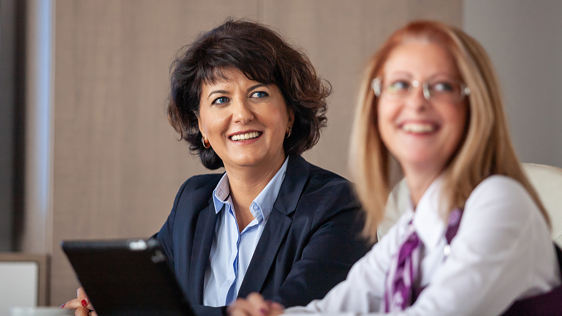 Two professionally dressed women sat at a table smiling with an ipad